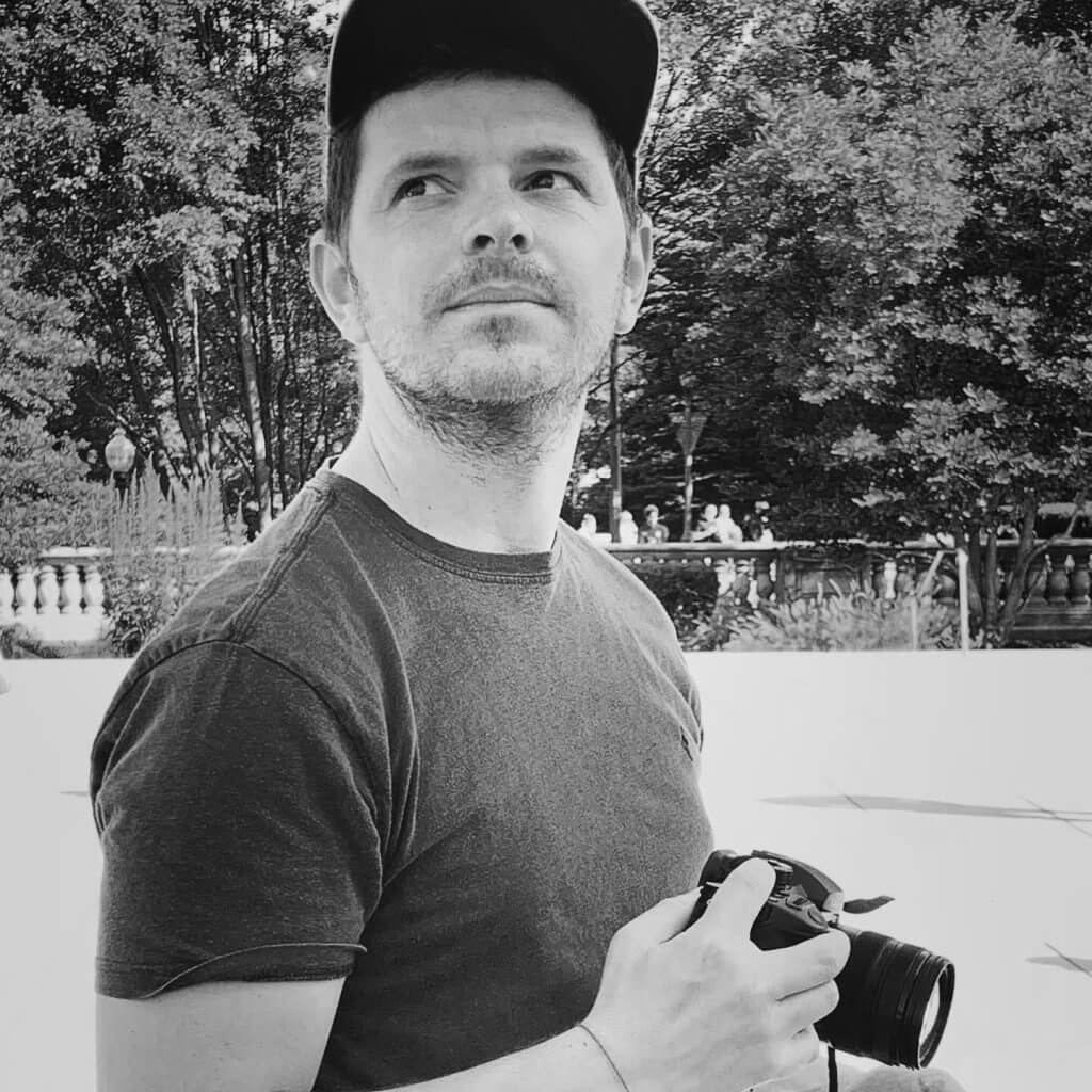 Mathieu Faure wears a T-shirt and cap and looks upward with his right hand holding a camera.