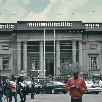 Long shot of a library in Nairobi, outside city life happens, cars are parked, a man with a jacket holds a cellphone in his hands, and two women walk behind him