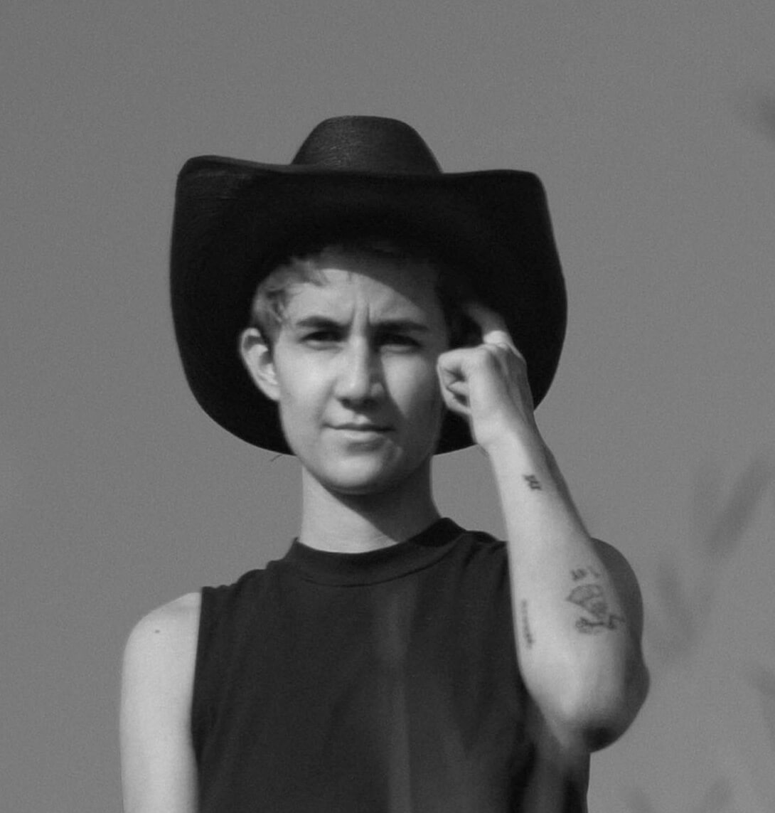 Portrait of Ana Rodriguez, wearing a sleeveless shirt, cowboy hat and touching the left side of her head with a finger