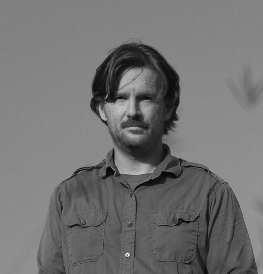 Photograph of a man with a buttoned shirt, medium hair and a mustache, looking at the camera