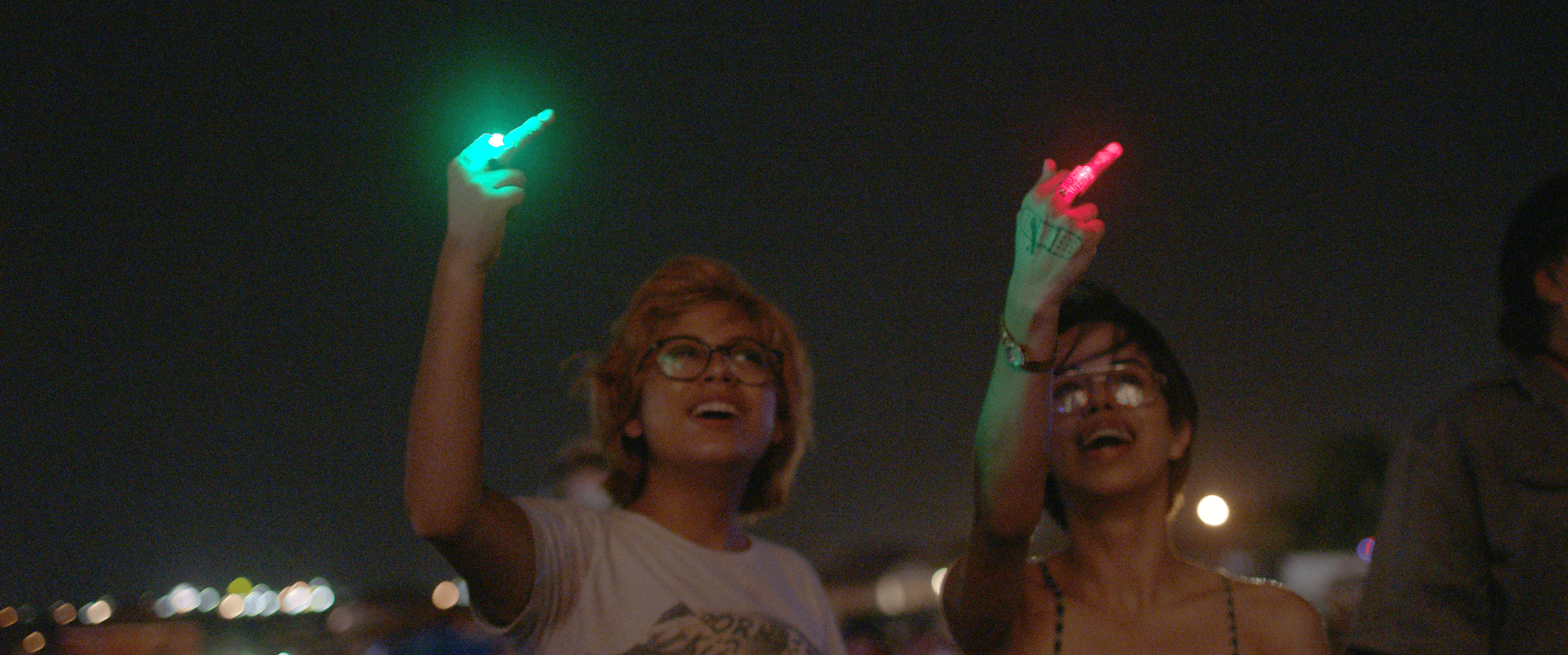 Silvia and Beba smile as they point their middle finger at the fire-work lit sky. There is a red light on Beba's middle finger, and a green light on Silvia's.