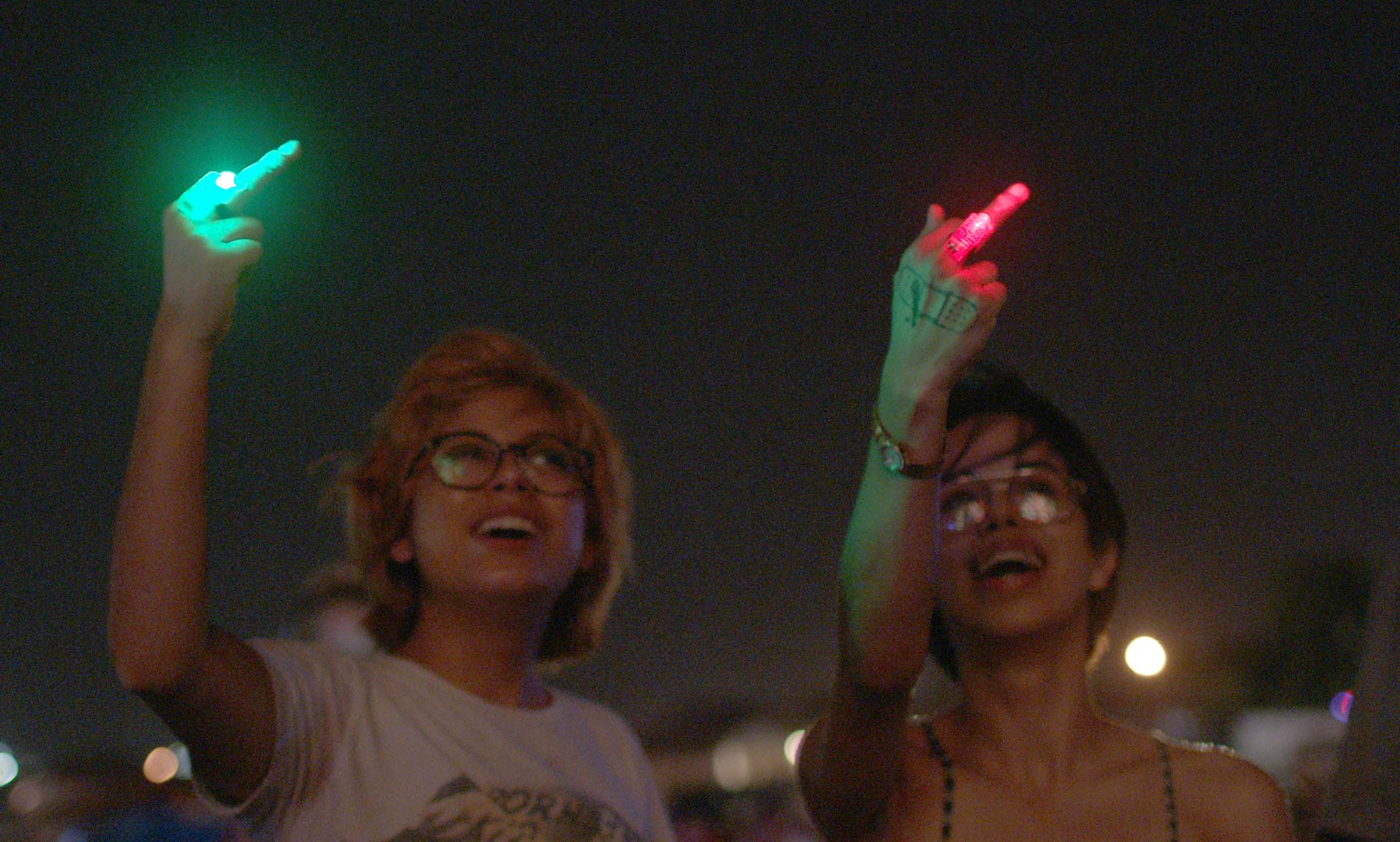 Silvia and Beba smile as they point their middle finger at the fire-work lit sky. There is a red light on Beba's middle finger, and a green light on Silvia's.