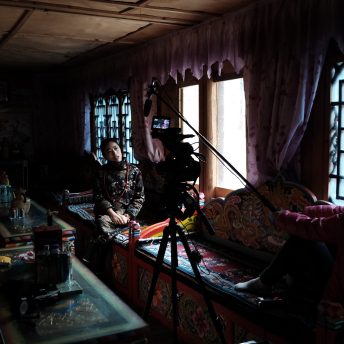 A young Tibetan woman is being interviewed in her home. The filmmaker holds the boom pole and the camera is visible in the foreground.