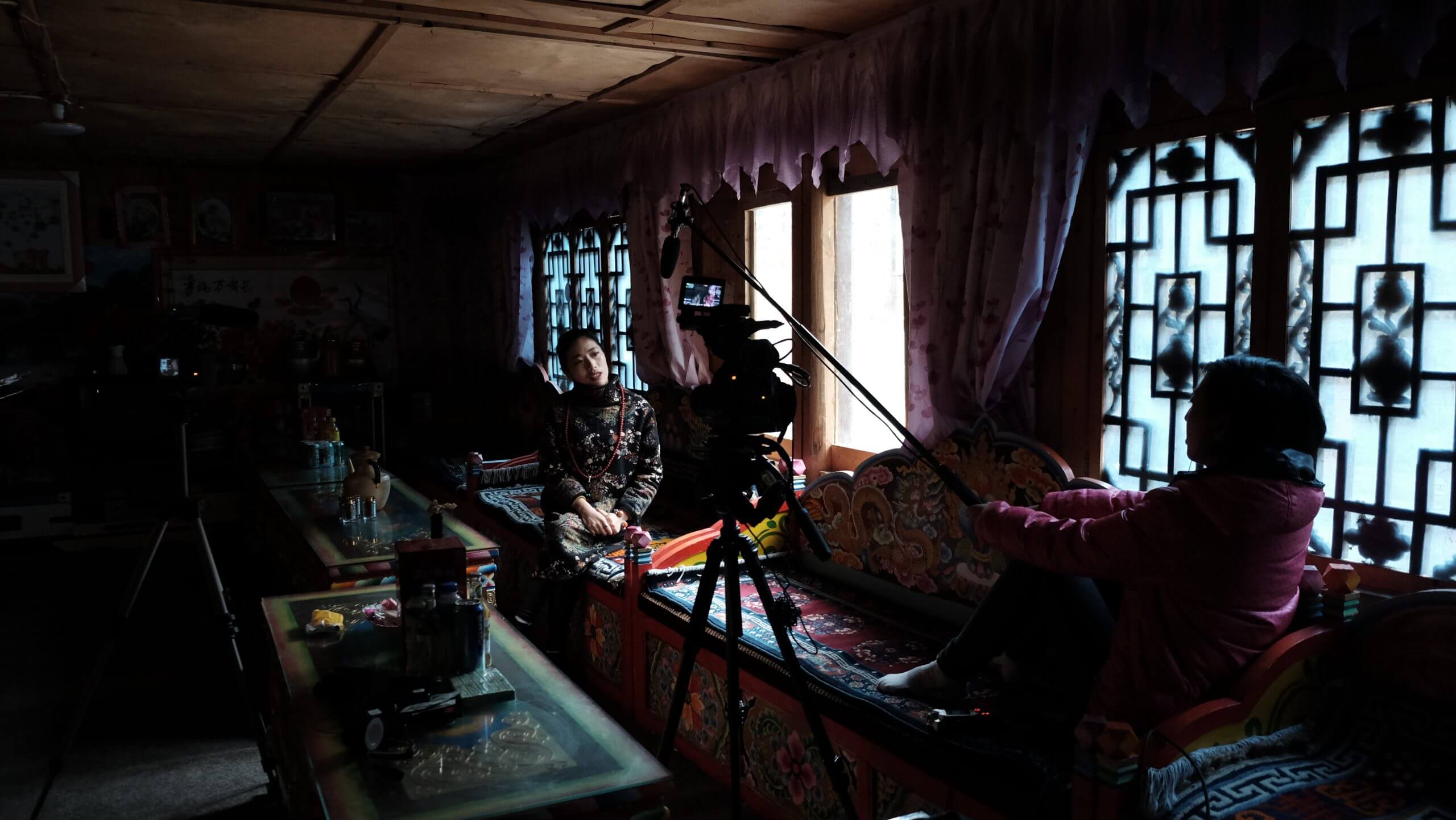 A young Tibetan woman is being interviewed in her home. The filmmaker holds the boom pole and the camera is visible in the foreground.