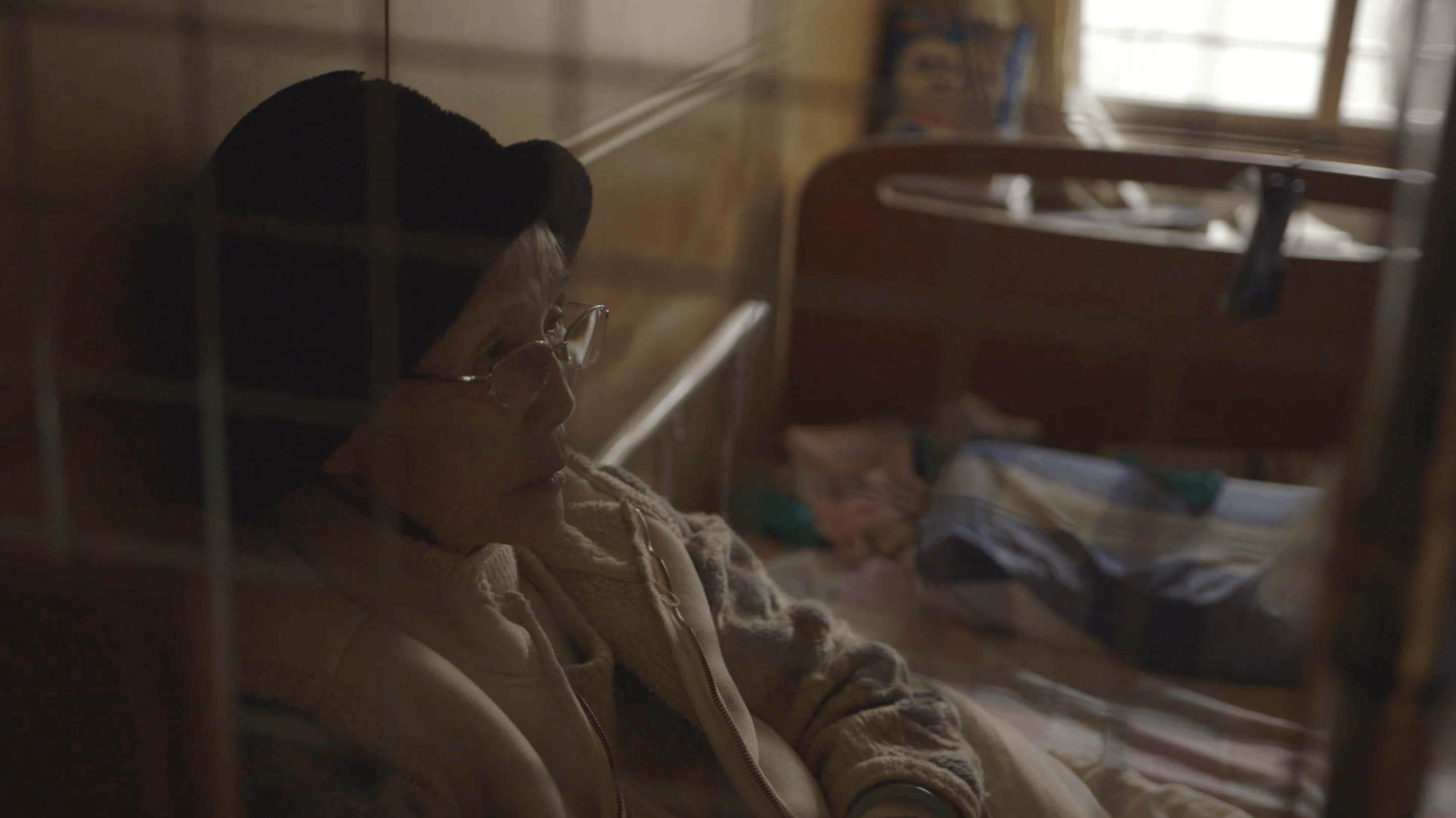 An older, Taiwanese woman with a black hat and glasses sits on her bed, behind a blurry grate in the foregound.