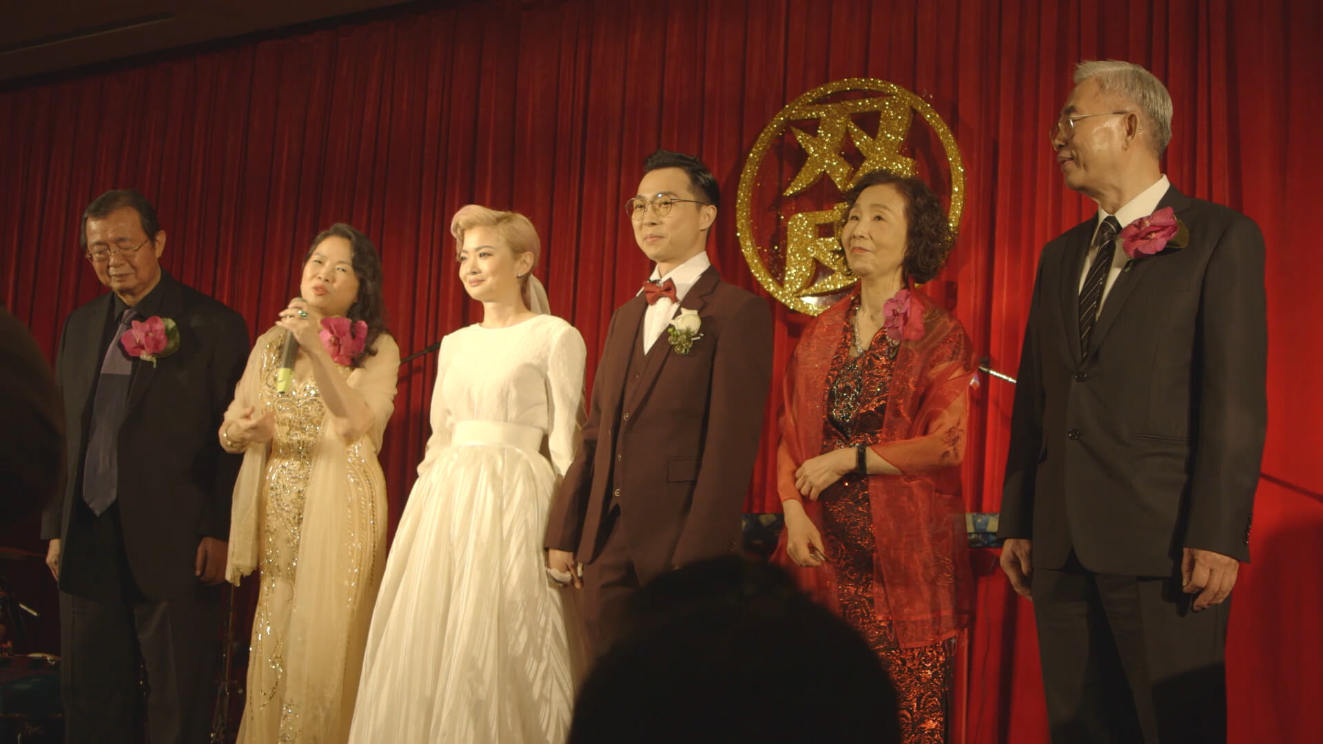 A young Taiwanese couple stands on a stage in front of a red curtain with their parents on either side of them. It's their wedding day.