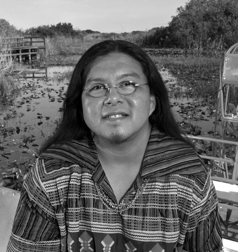 Houston, a two spirit member of the Miccosukee Tribe, has long dark hair, glasses, and is wearing a Miccosukee patchwork top