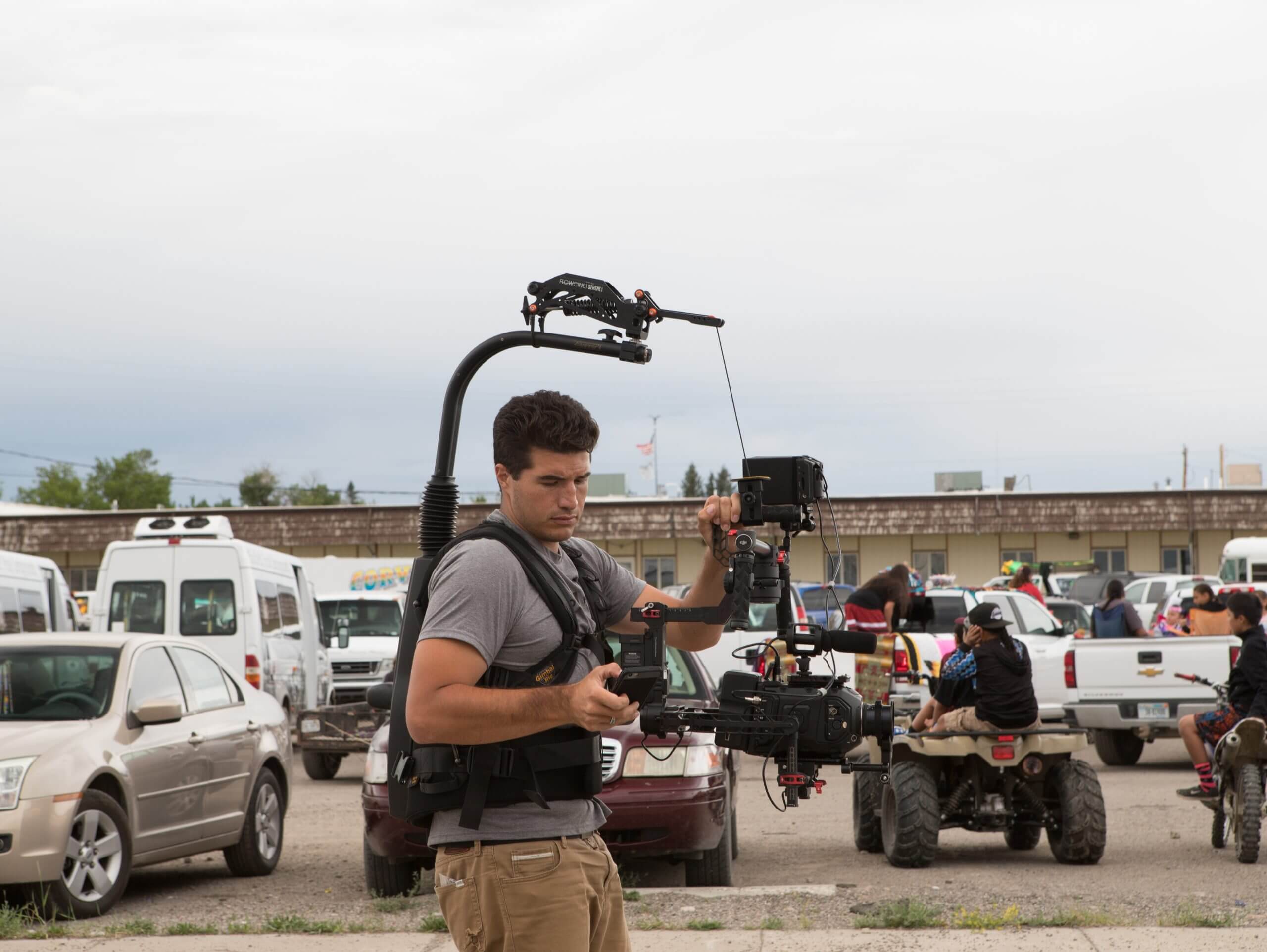 A cameraperson holds a camera harness, in the background cars and trucks are parked, there is a motorcycle with children looking at something