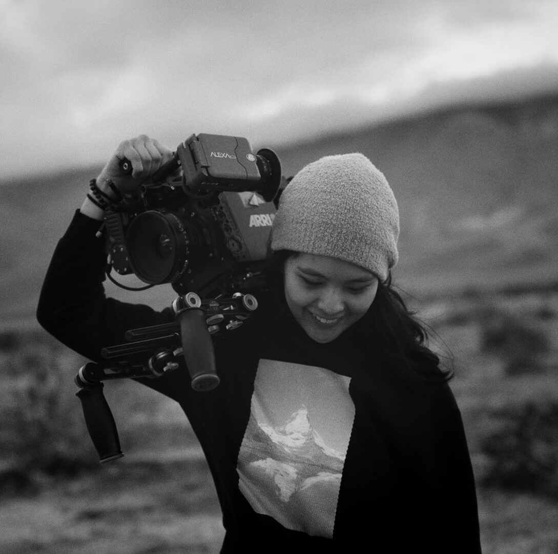 Portrait of a woman looking at the ground while holding a camera on her right shoulder, she wears a hat and a long sleeve shirt