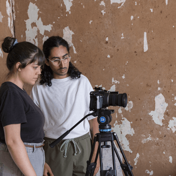 Gabriela and Picho stand behind the camera, they are in front of a wall with paint peeling off