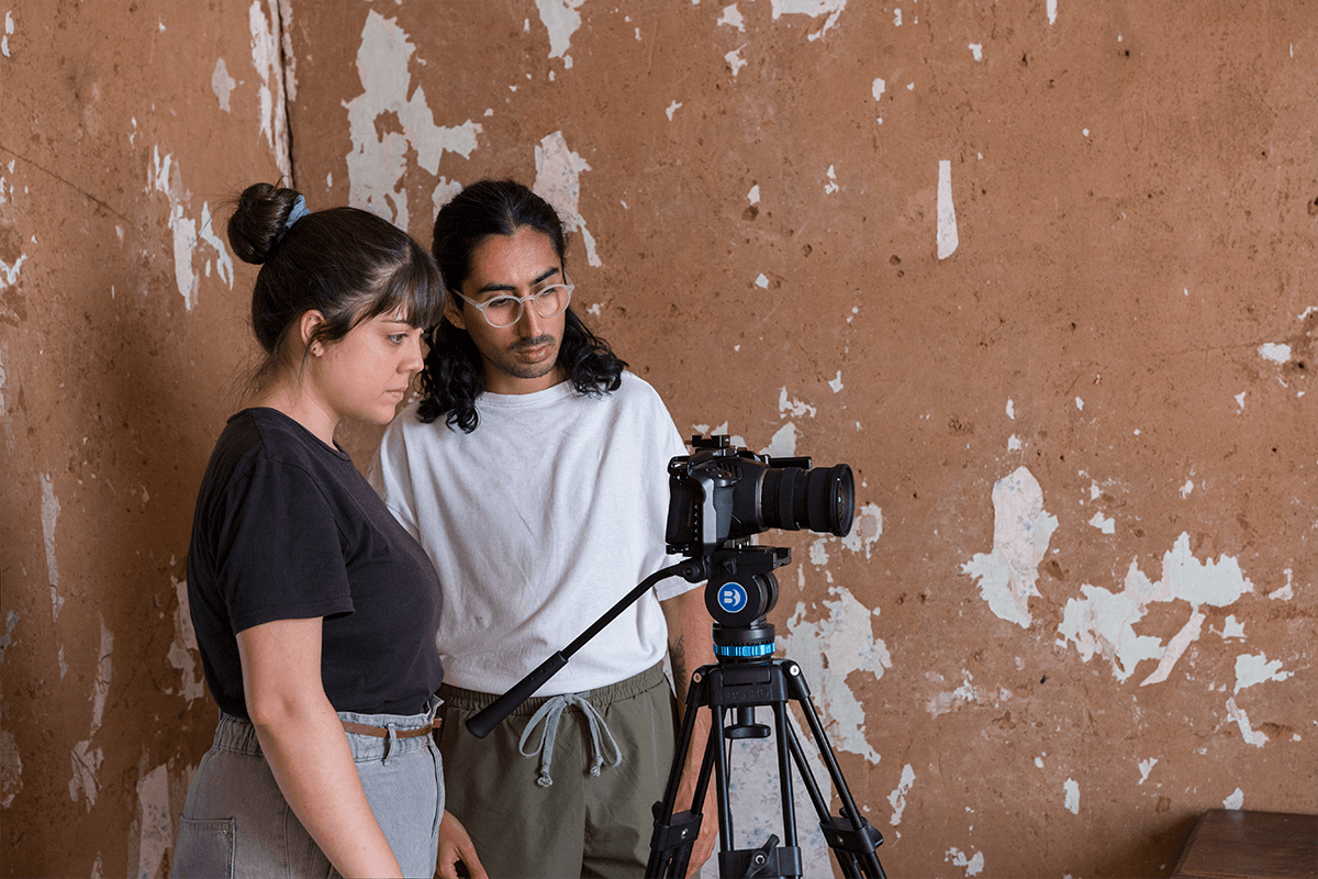 Gabriela and Picho stand behind the camera, they are in front of a wall with paint peeling off