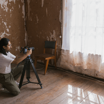 Picho is on his knees working with the camera, in front of him there is a window with a white curtain
