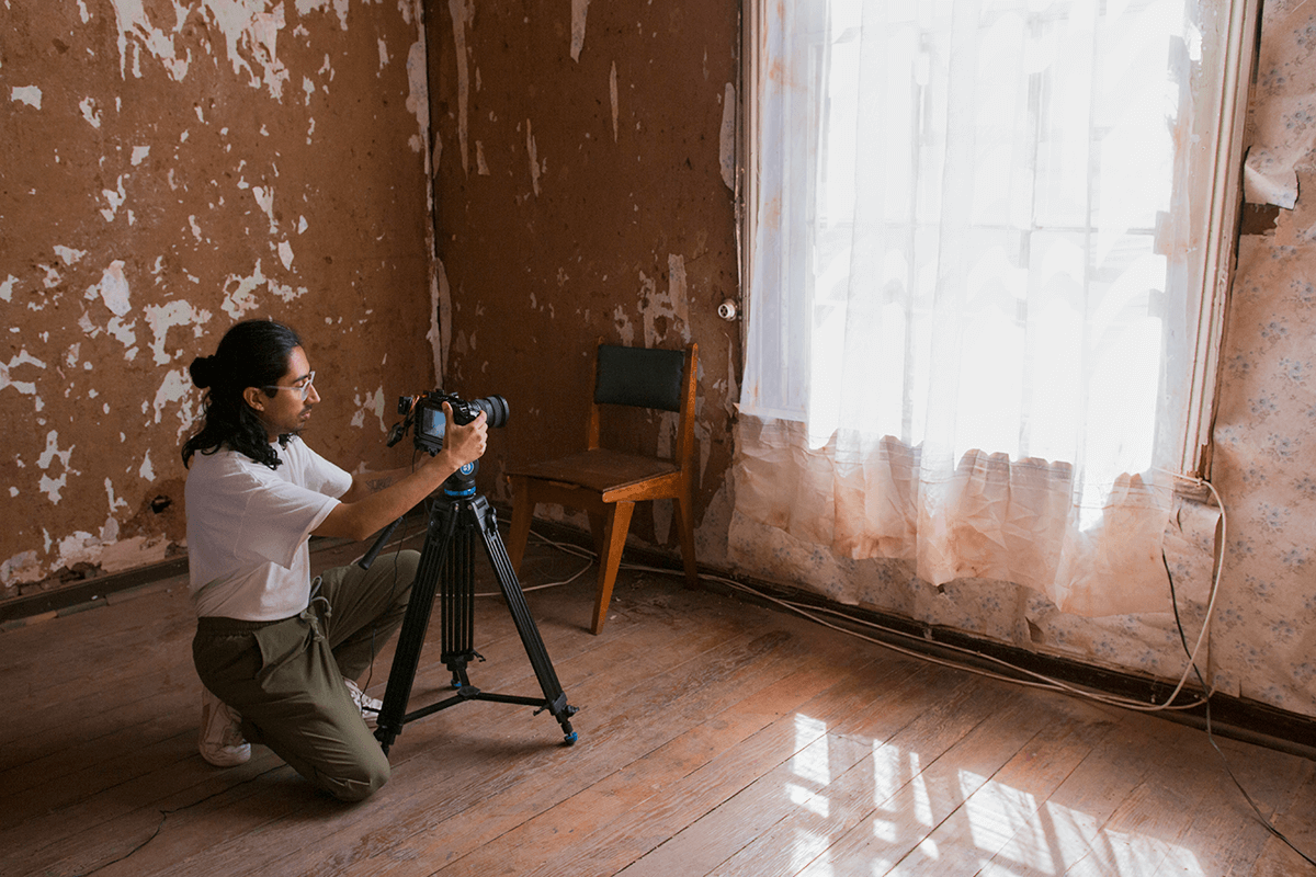 Picho is on his knees working with the camera, in front of him there is a window with a white curtain