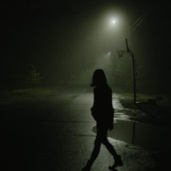A silhouette of a young woman is walking across an empty and foggy suburban street at night.