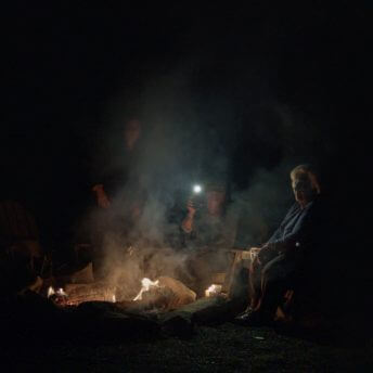 Three woman are gathered around a campfire at night. The grandmother is looking off into the distance, the mother is holding a flashlight, and the daughter's face is obscured in smoke.