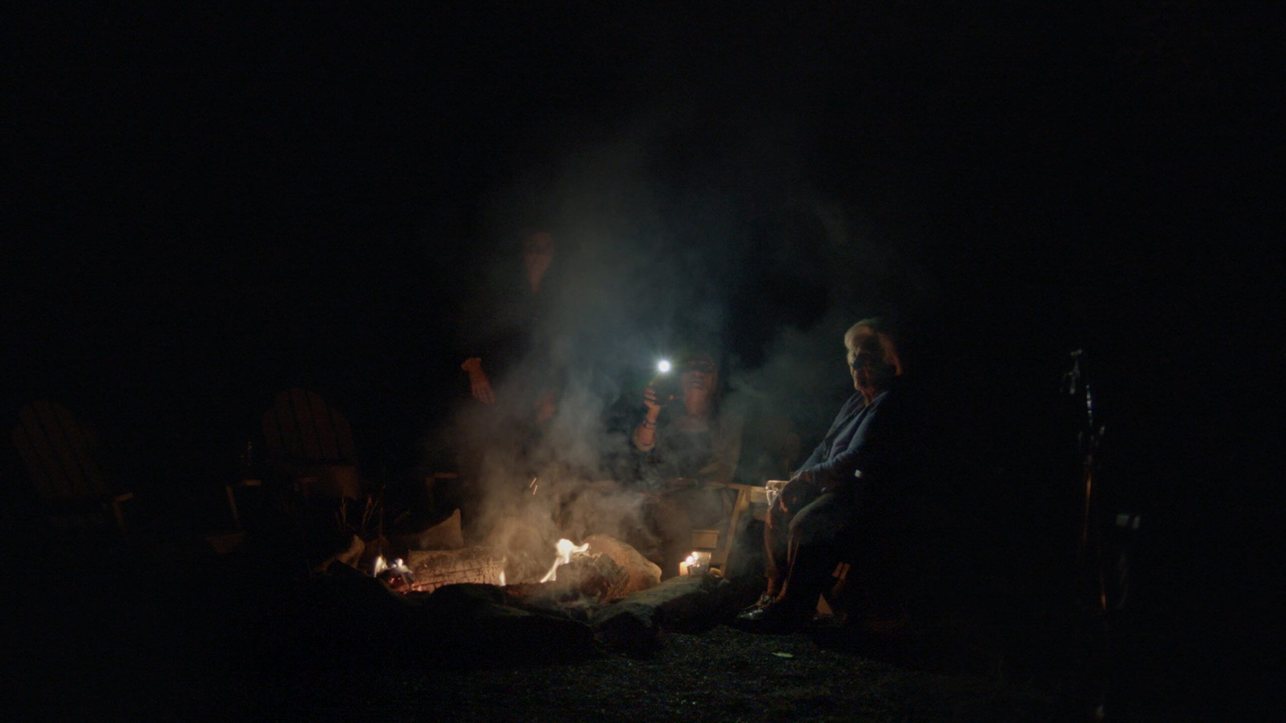 Three woman are gathered around a campfire at night. The grandmother is looking off into the distance, the mother is holding a flashlight, and the daughter's face is obscured in smoke.