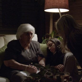 Three generations of women are gathered under a lamp on a large porch at night. The grandmother is sitting on a couch telling a story, the mother is leaning in to looking at something the grandmother is drawing, and the daughter has her eyes closed.