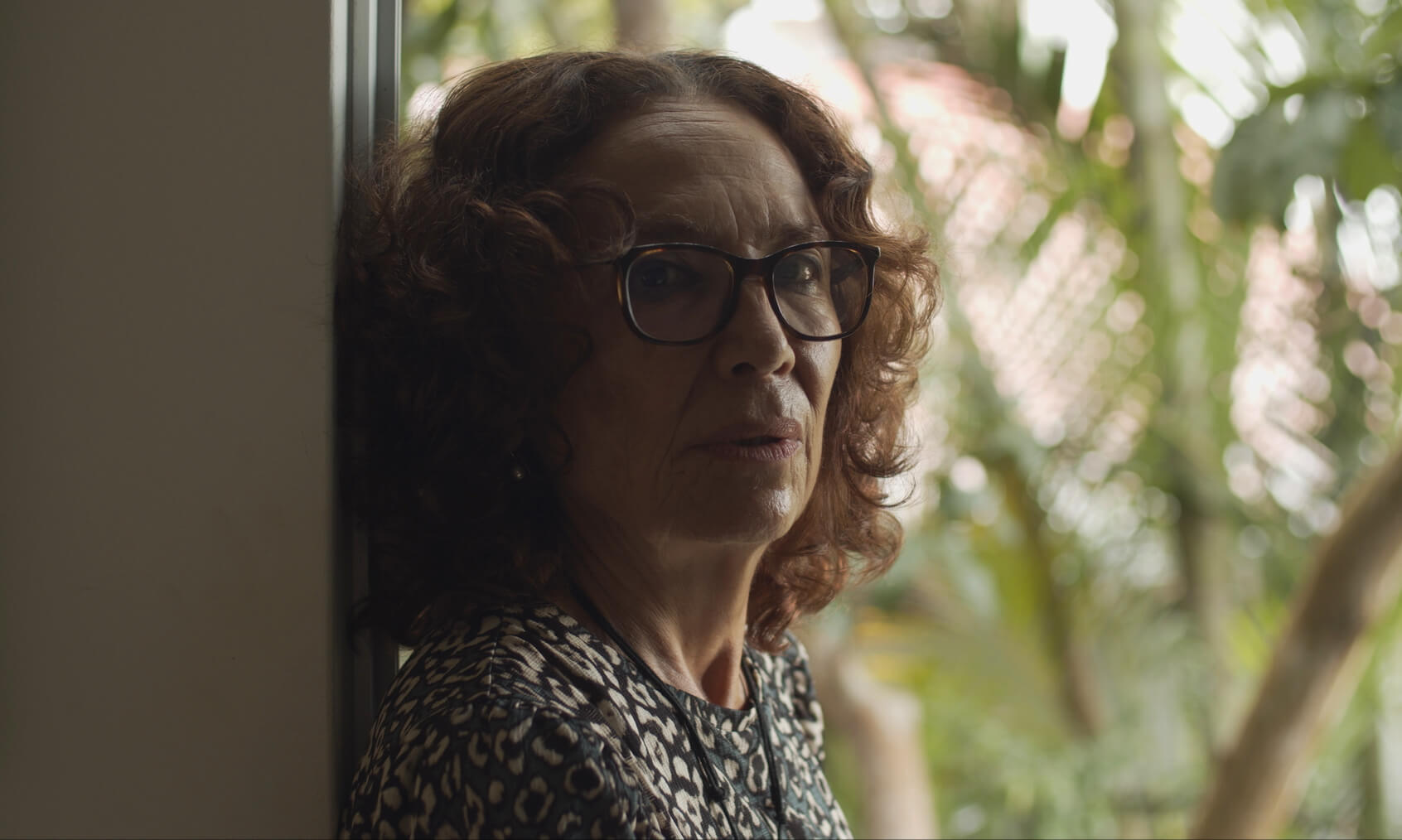 Close up of a woman leaning against the frame of a door, she has curly shoulder length hair and wears glasses