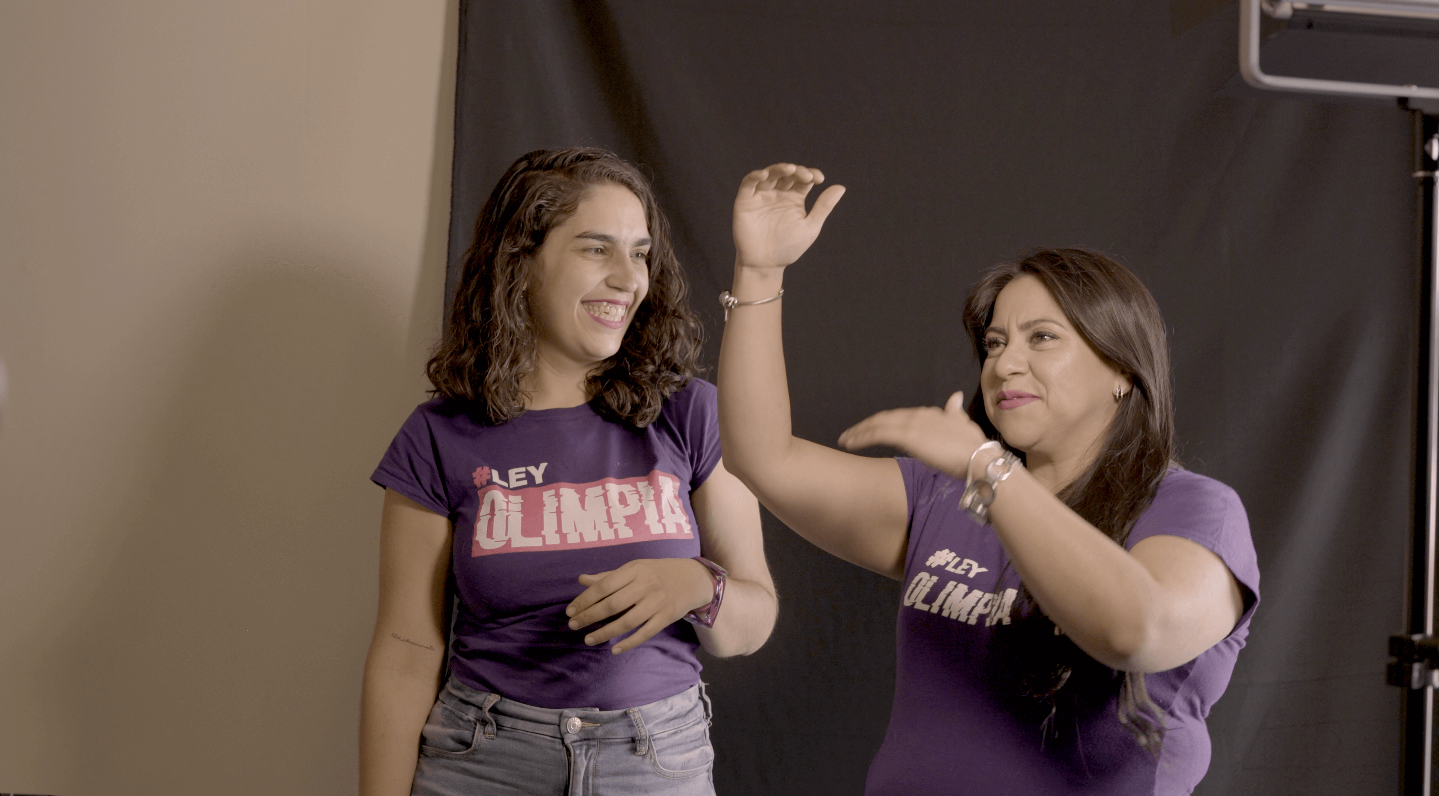 Olimpia Coral and Director Indira Cato laughing during a photoshoot. Both wear a purple "Olimpia Law" t-shirt.