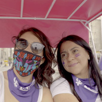 Olimpia Coral and Director Indira Cato taking a selfie at the International Women's Day protest. Indira wears sunglasses and a face mask, Olimpia has her face painted with purple marker, both wear "Olimpia Law" handkerchiefs.