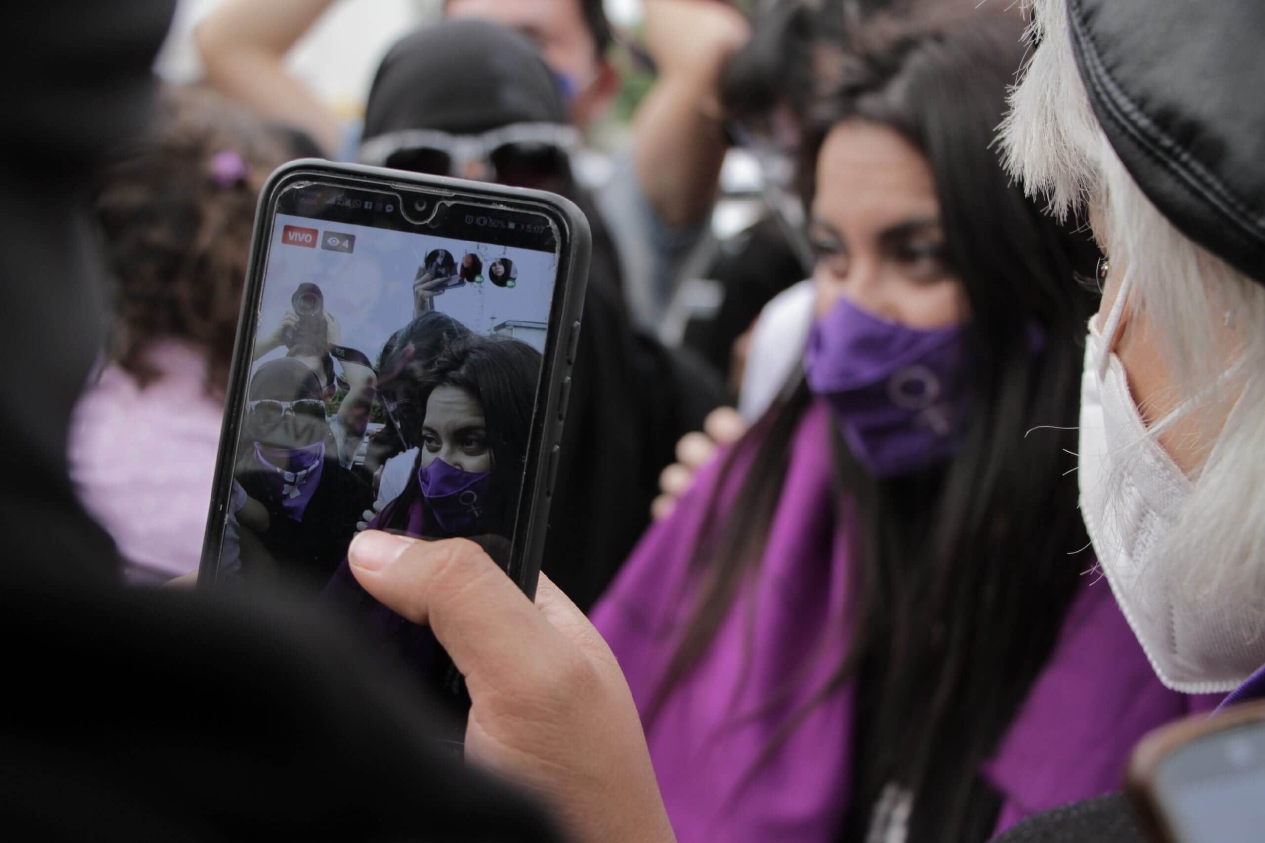A phone recording Olimpia Coral and others outside the Chamber of Deputies before the Olimpia Law's approval. Olimpia wears a purple mask and a purple flag over her shoulders.