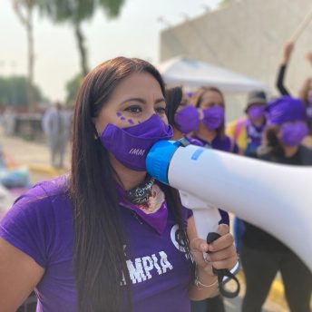 Olimpia Coral holding a megaphone outside the Mexico City Chamber of Diputies during the Olimpia Law's approval. She wears a purple face mask and an "Olimpia Law" t-shirt.