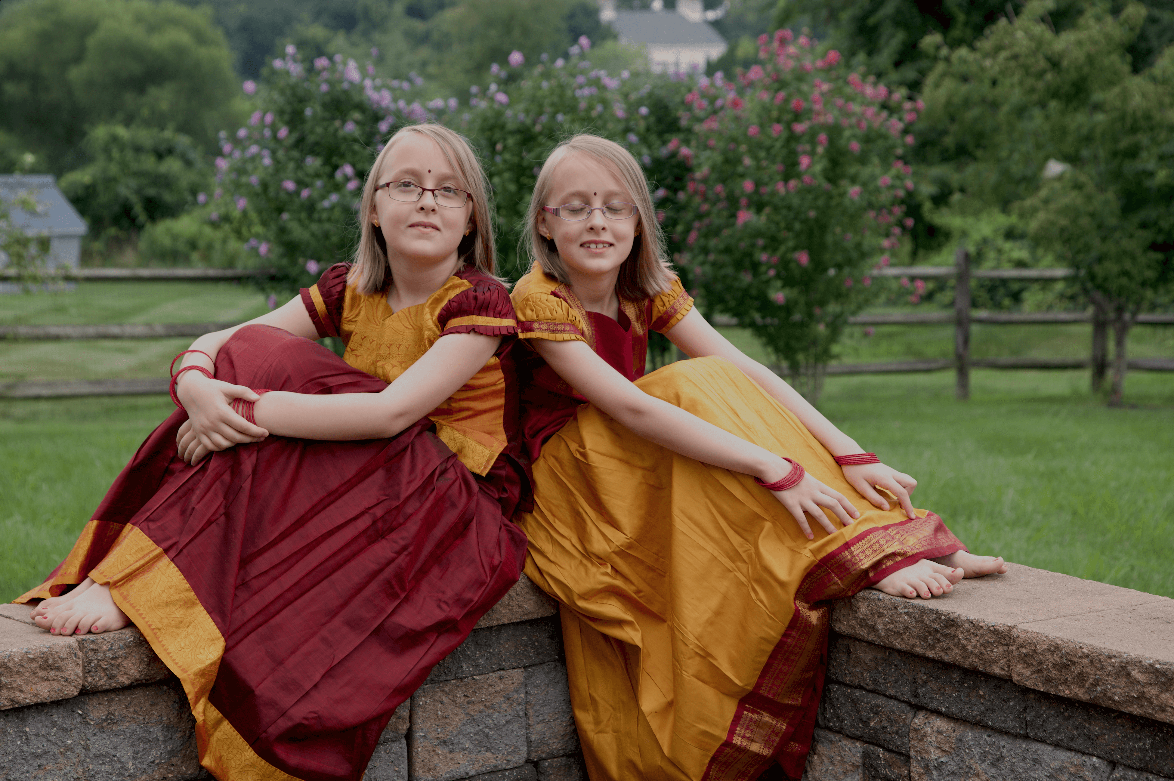 Fraternal white twins Anjali and Meghna dressed in traditional south Indian Kancheepuram silk skirts and blouses in red and mustard colors are sitting and leaning against each other. Meghna's arms are crossed around her knee and she is looking at the camera. Anjali is smiling with her eyes closed and her arms are stretched out and resting on her legs. Both are wearing spectacles, red dots on their forehead and red bangles, and their nails are painted red. They have shoulder length blonde straight hair.