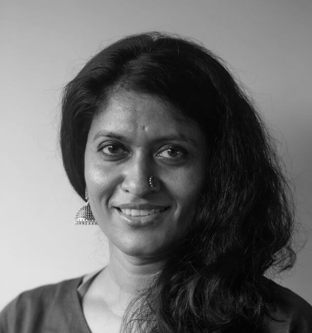 A warm color portrait of Chithra Jeyaram, a South Asian filmmaker, smiling directly at the camera. She's wearing a navy blue top, a stone-studded nose ring, and large semi-dome-shaped silver earrings. Her shoulder-length dark brown hair is gathered on her left shoulder.