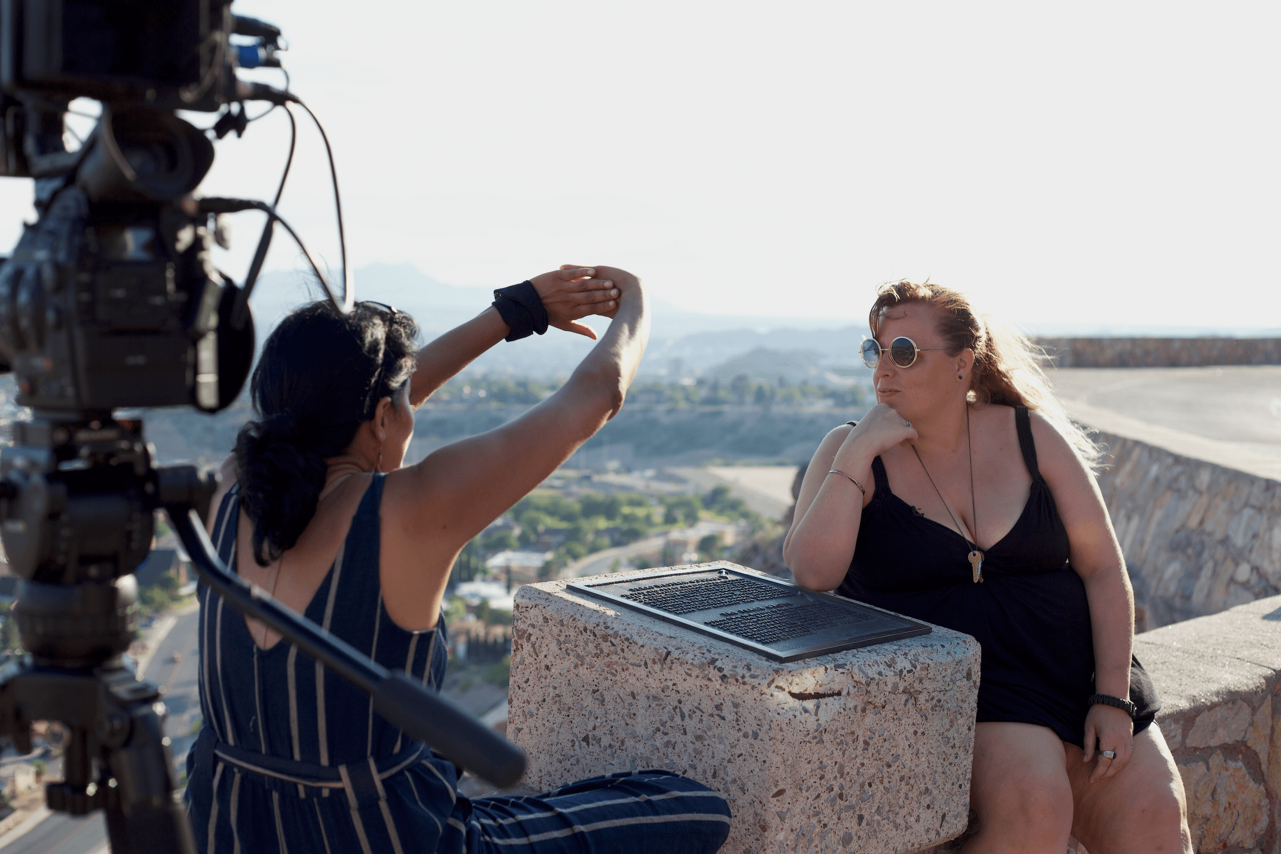Chithra Jeyaram, a south Asian filmmaker, and Brandy Stein, a white woman, are sitting facing each other at a scenic outlook at magic hour. Chithra is facing away from the camera with her arms outstretched to block the sun while in conversation with Brandy, who is facing the camera. Brandy has sunglasses and her right elbow is resting on a plaque with the hand under her chin. In the foreground to the left, is a camera on a tripod, both out of focus.