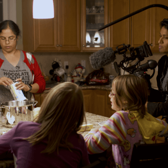 In a suburban kitchen at night, Lakshmi a South Asian woman is unwrapping butter into a stainless steel container, across her at the island are seated her adopted fraternal twins Anjali and Meghna. They are white with blonde hair and are watching their mother at work. To the right, Chithra Jeyaram, a south Asian filmmaker with a camera rig is filming Lakshmi.
