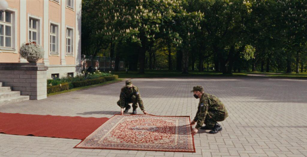 Two soldiers on their knees lay out a rectangular carpet outside a big house