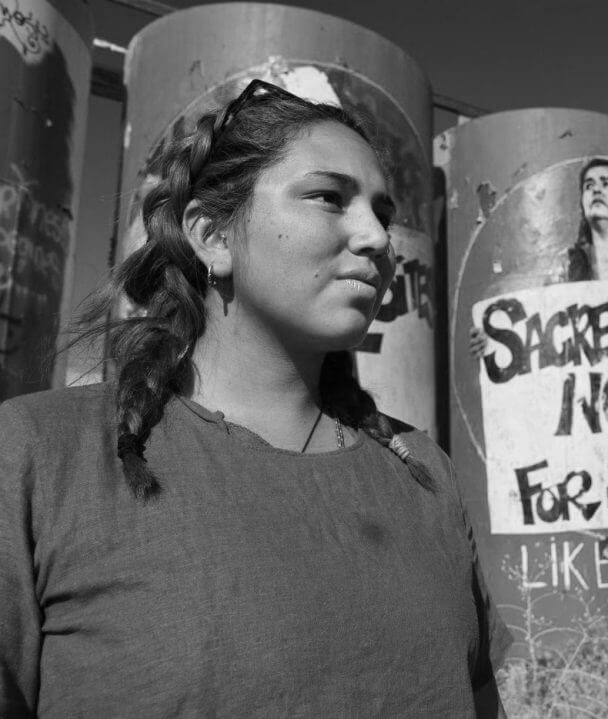 Portrait of Ivey Camille Manybeads Tso. Ivey looks away from the camera, with braided hair and a t-shirt. In the back there are columns with posters pasted that protest the sale of Native land.