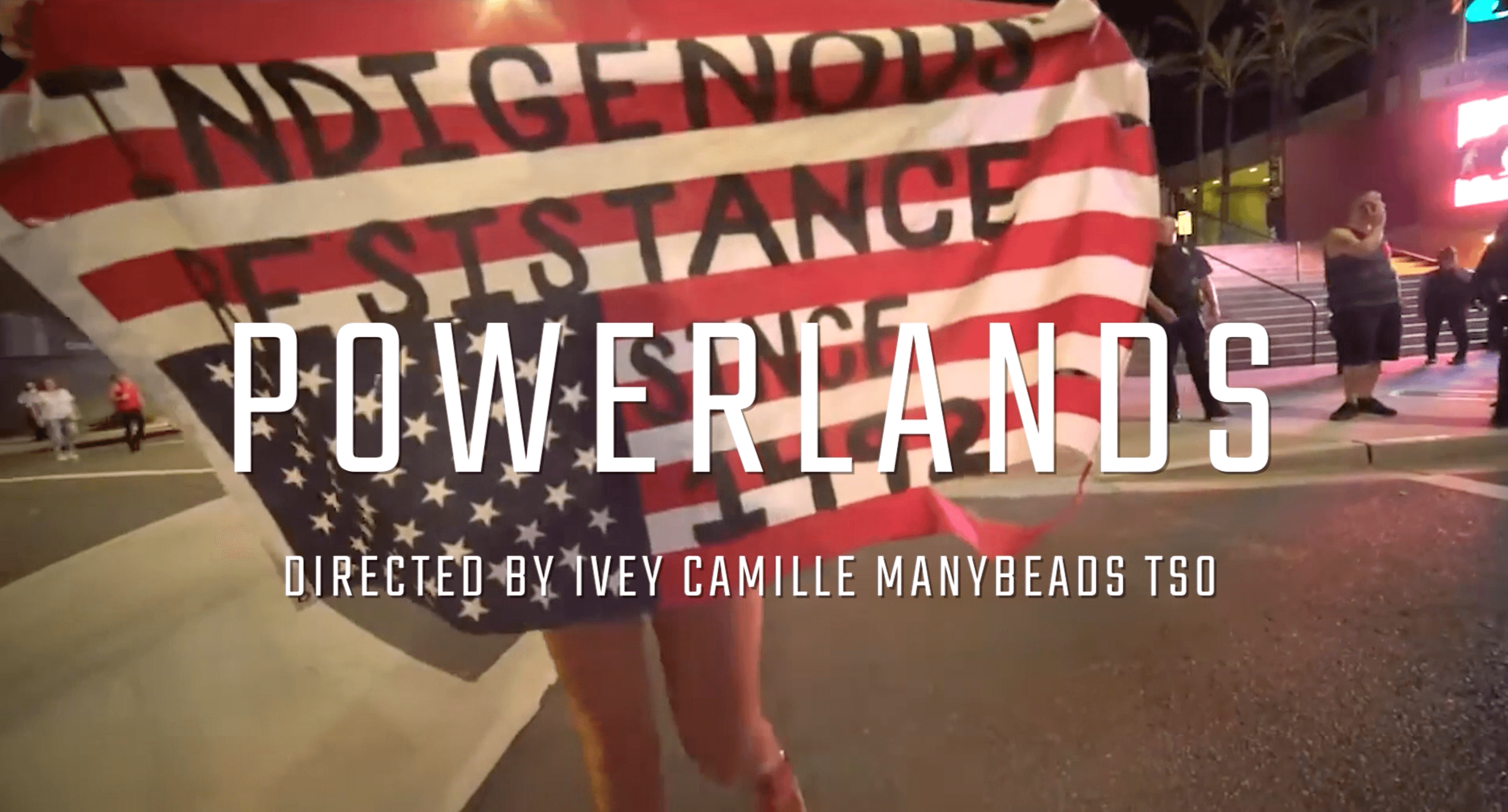 Still from the film Powerlands, a person holds a US flag that is upside down and says Indigenous Resistance since 1492, on top of it there is the title of the film and the name of the director