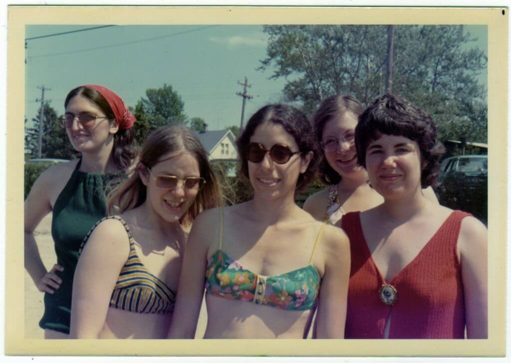 A photograph of 5 women wearing swimsuits and shades, in a sunny day in the'70s.