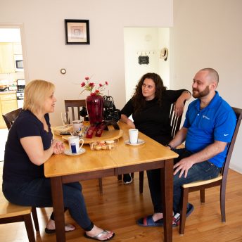 Three people are sat in a dining room during the day. On the left thee is a woman with short blond hair and wearing black t-shirt and pants, in front of her there are a woman with long hair and black long sleeve shirt, and a man with beard and a blue polo shirt, bot look at the blond woman. In the table there are cookies, coffee mugs, flowers in a vase and a camera. In the background you can see the entrance to a kitchen.