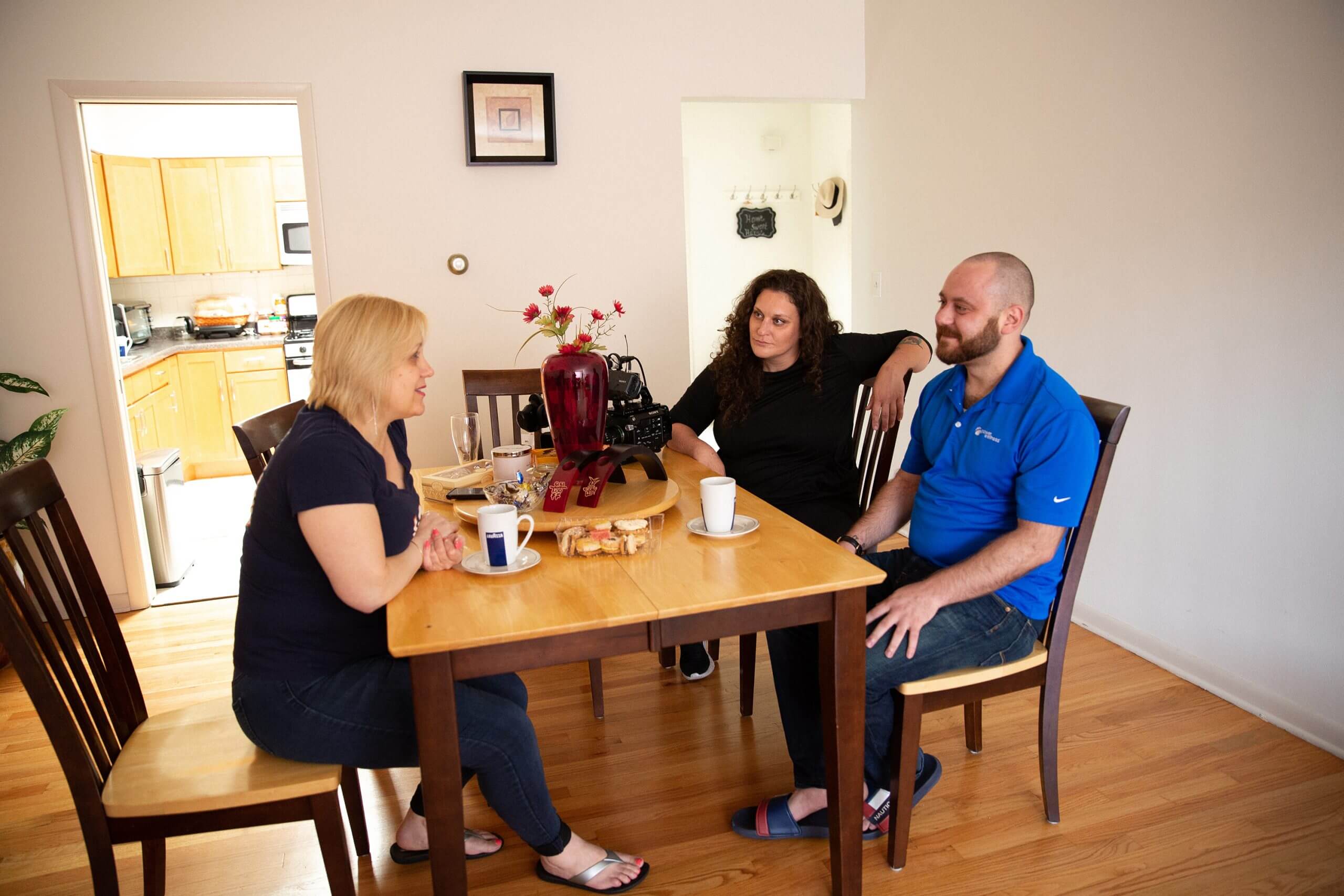 Three people are sat in a dining room during the day. On the left thee is a woman with short blond hair and wearing black t-shirt and pants, in front of her there are a woman with long hair and black long sleeve shirt, and a man with beard and a blue polo shirt, bot look at the blond woman. In the table there are cookies, coffee mugs, flowers in a vase and a camera. In the background you can see the entrance to a kitchen.