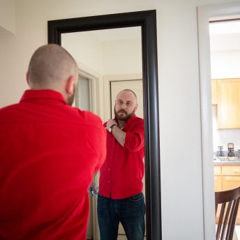 Over shoulder shot of a bald man with a bear, wearing a red shirt looking himself in front of a mirror while resting his arm in front on his shoulder