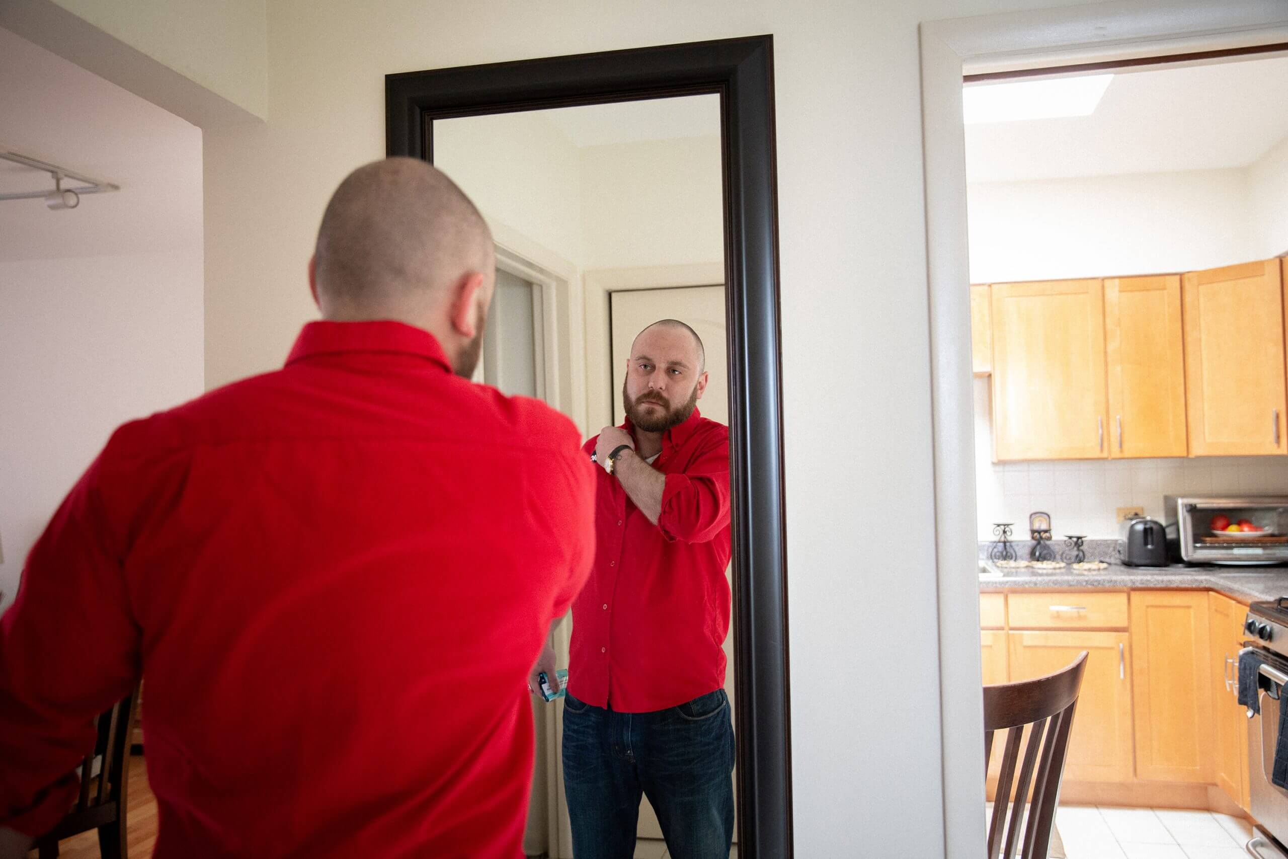 Over shoulder shot of a bald man with a bear, wearing a red shirt looking himself in front of a mirror while resting his arm in front on his shoulder