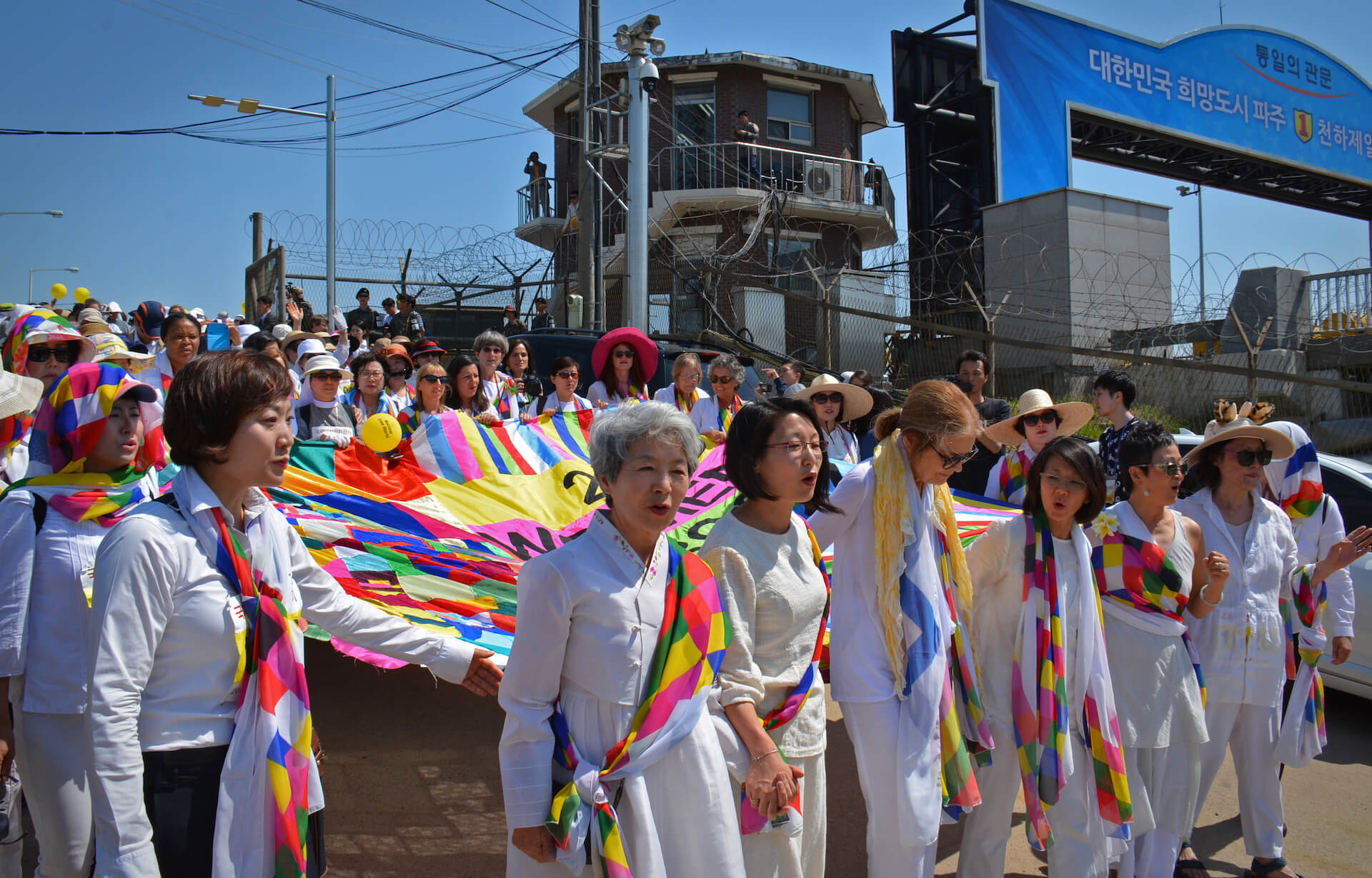 A group of many women walking dressed in white with a colorful SCARF. They hold a colorful banner. The walk past a watch tower and a wired fence.