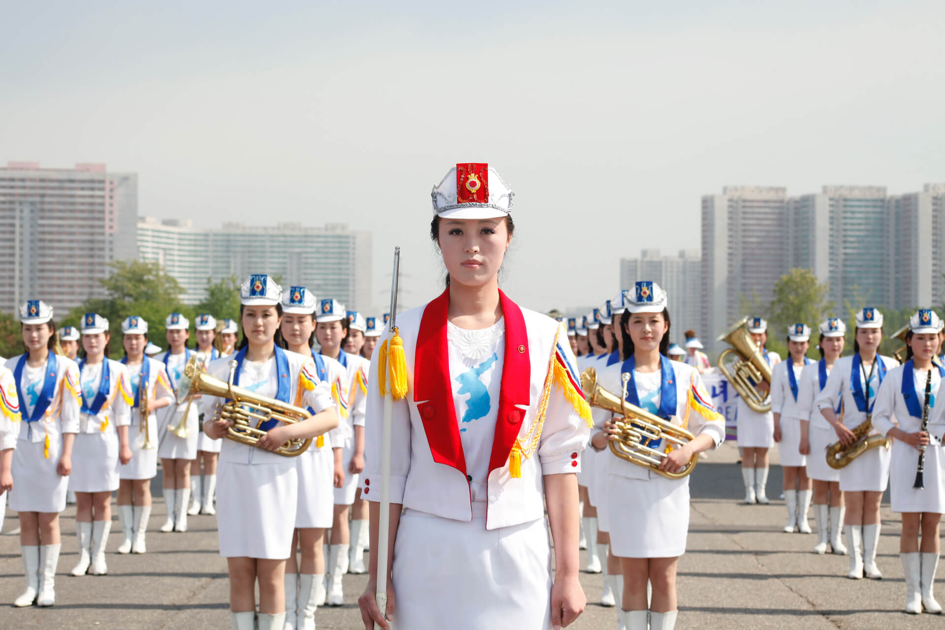 Medium shot of a Korean woman dressed in white, with the shape of Korea in her t-shirt, on the back there are more women in lines, dressed in white as well and some of them hold instruments.