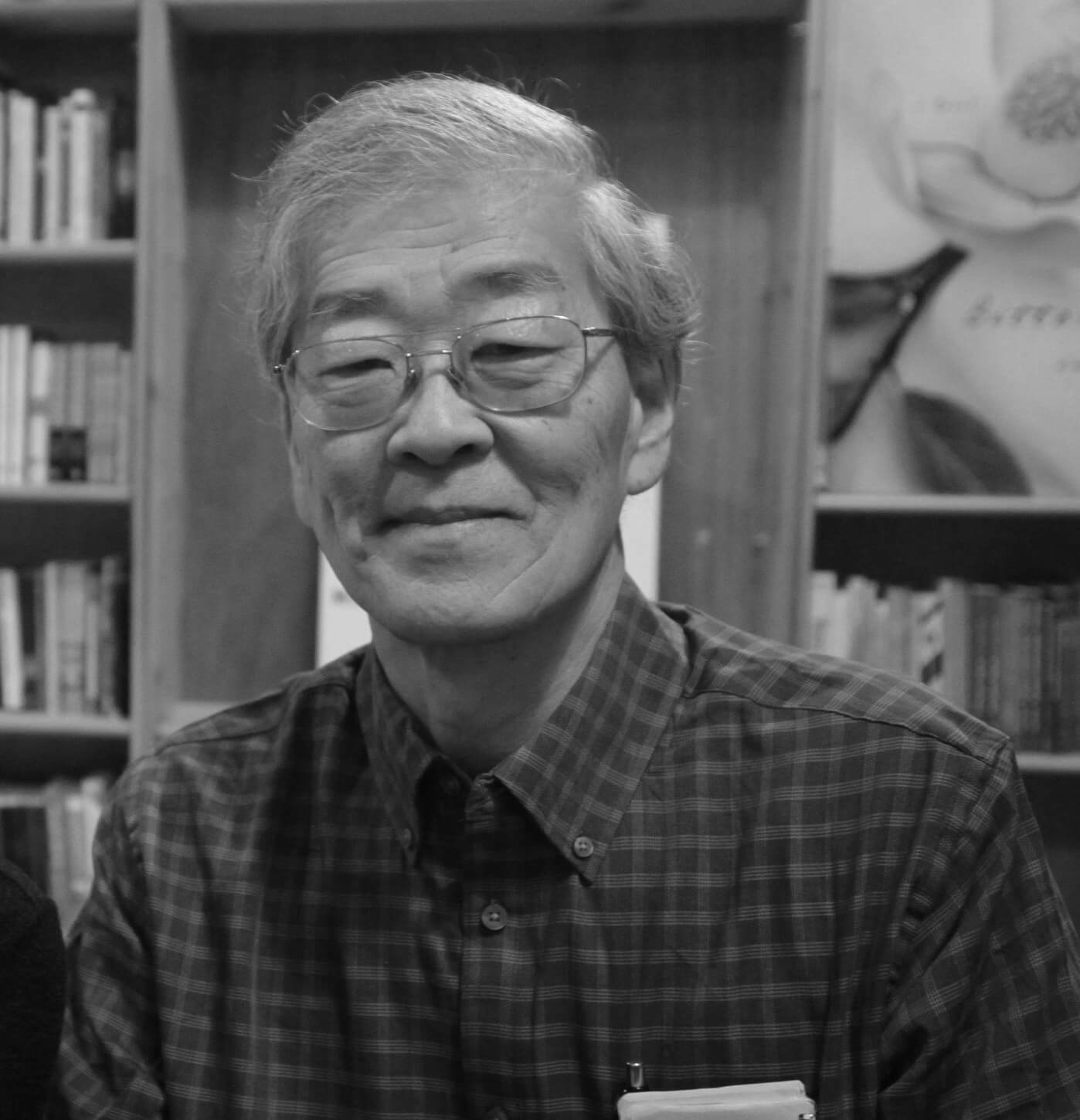 Portrait of Ramsay Liem, he wears a checkered shirt, and thin frames. He smiles at the camera. In the background there is a bookshelf and a painting.