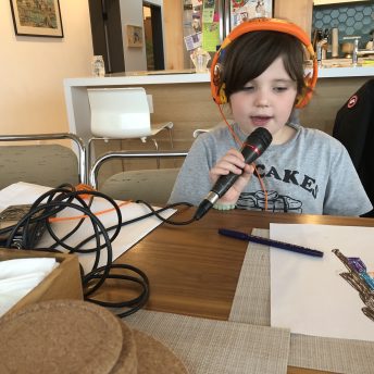 A young girl wears headphones and talks through a microphone connected to a tascam, while watching a drawing on the table.