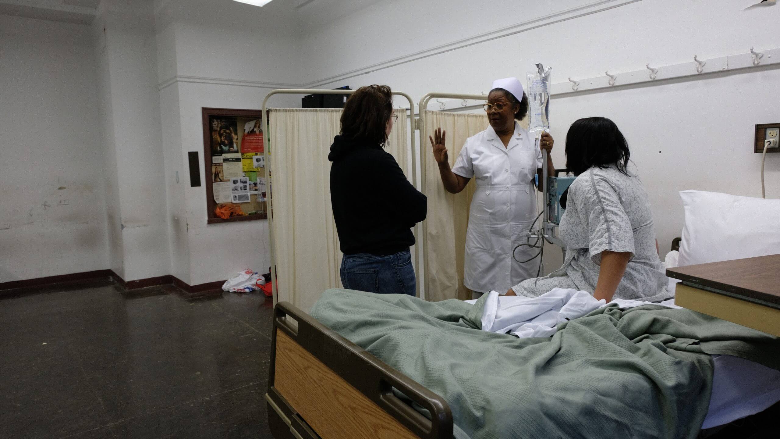 Director Emma Francis-Snyder talking to an actress dressed as a nurse, and an actress dressed as a patient sat on a bed