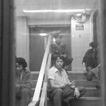 Taken from inside of a room, a shot of four actors dressed as members of the Young Lords Party sat on a stair