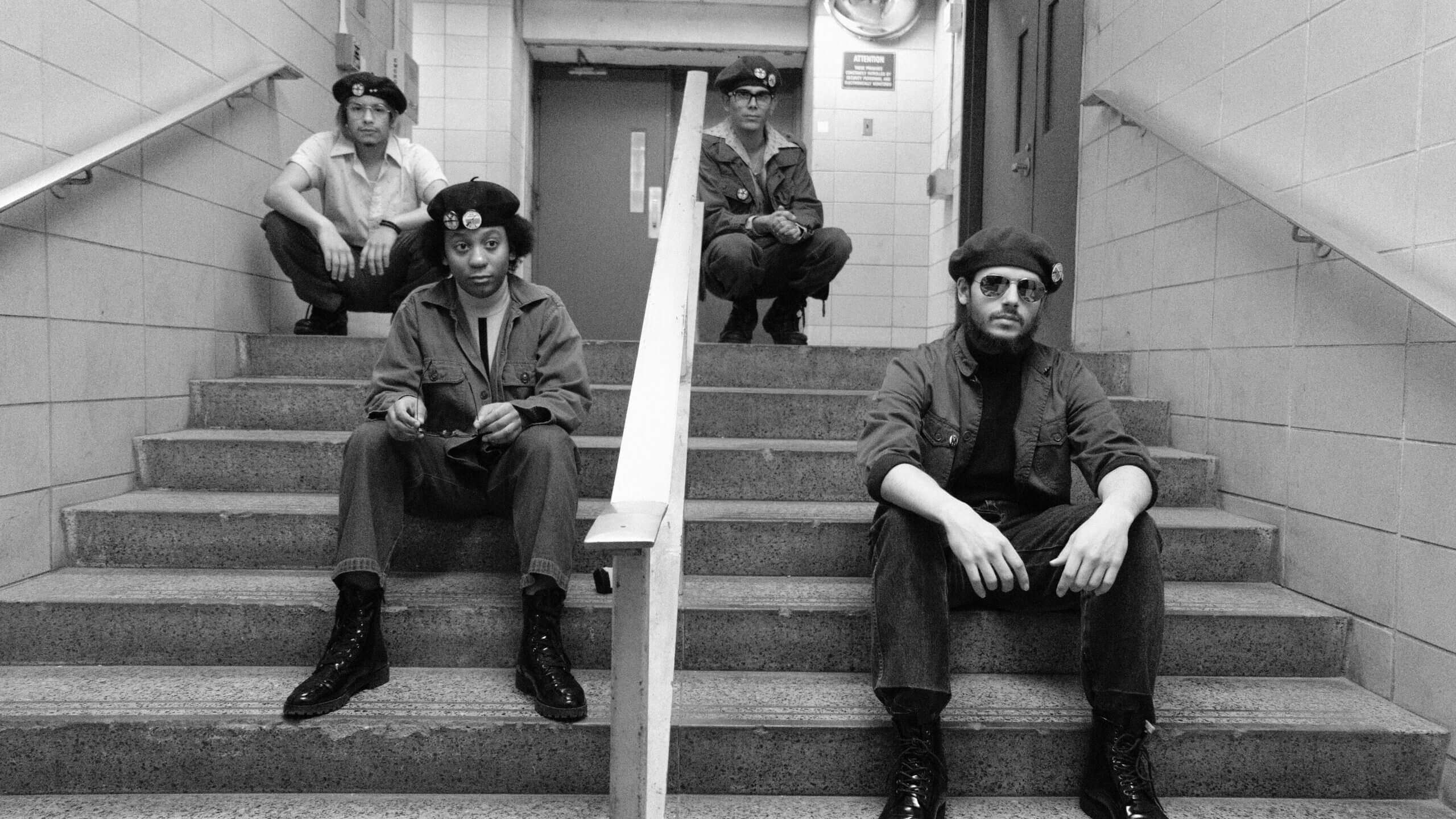Low angle shot of four actors dressed as members of the Young Lords Party sat on a stair