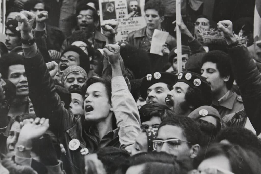 A photography of a demonstration, there are men an women, some of them wear berets, some raise their fists up, some hold signs, many are screaming something