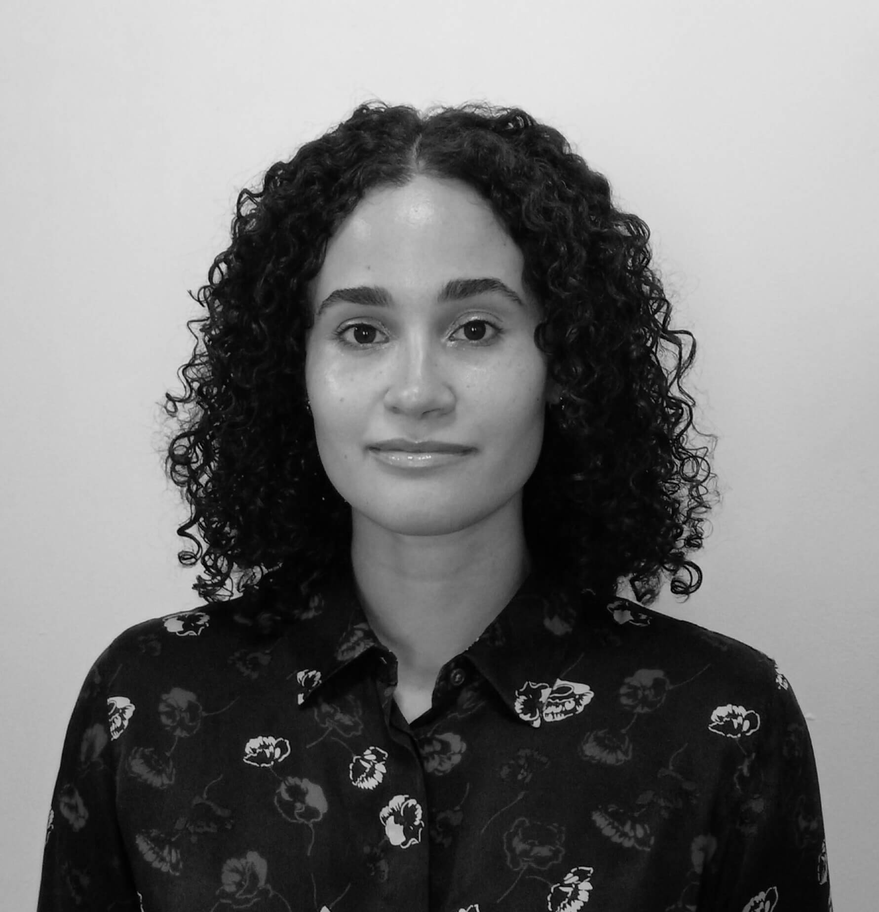 Allison Villegas Roman smiles at the camera. She has curly hair, and wears a buttoned shirt with flowers. Portrait in black and white.