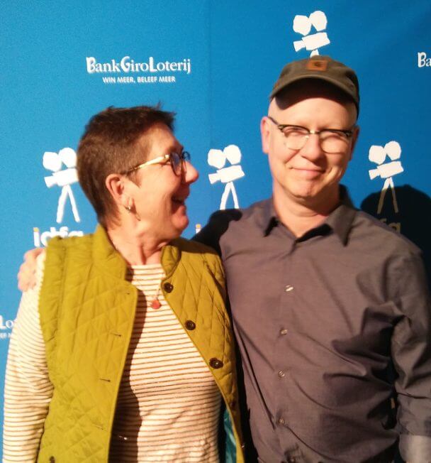 Julia Reichert looks at Steven Bognar and smiles. Steven Bognar looks at the camera and smiles. Both of them are in front of a blue photo call with white cameras.