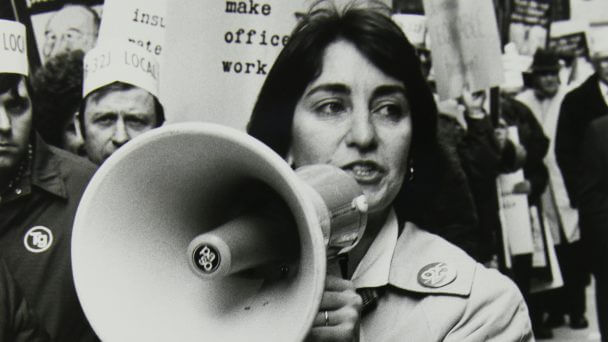 Still from 9to5: The Story of a Movement. White and black photograph of a person with a speaker close to their mouth. In the back, there are people with posters.