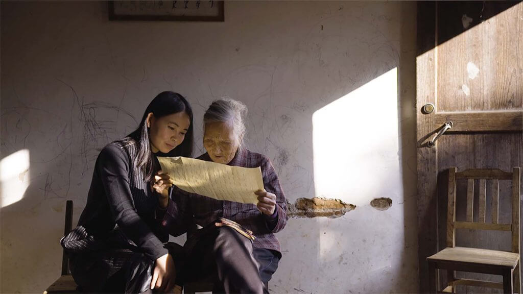 Two Chinese women sit next to each other. The young wan inclines her head to read what the elderly woman has in her hands: a yellow paper. The sunlight illuminates part of the room.